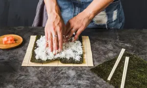 Dłonie rozprowadzają ryż haruka na nori, przygotowując się do zwinięcia sushi. Obok leżą pałeczki i marynowany imbir.