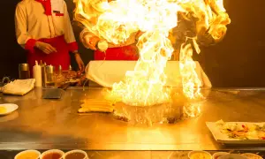 Szef kuchni tworzy spektakularny pokaz ognia podczas przygotowywania yakiniku. Płomienie unoszą się nad płytą grillową, a obok leżą kawałki mięsa.