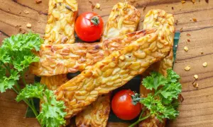 Usmażony tempeh, czyli co to jest? Złociste kawałki na drewnianej desce, ozdobione pomidorkami i natką pietruszki.