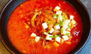 Pikantna zupa kimchi z kurczakiem i kostkami tofu, udekorowana szczypiorkiem.