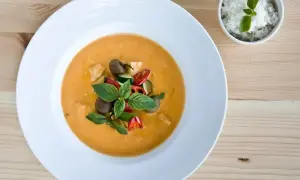 Aksamitny **thai red curry** z kurczakiem, cukinią i pomidorkami, udekorowany bazylią. Obok miseczka ryżu.