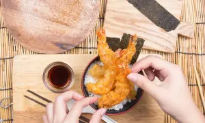 Ręce sięgają po chrupiące krewetki w panierce tempura, ułożone na ryżu z nori.