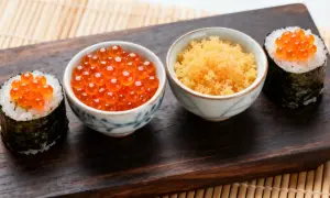 Dwa sushi z ikrą łososia i miseczki z ikrą masago i ikrą łososia na drewnianej desce.