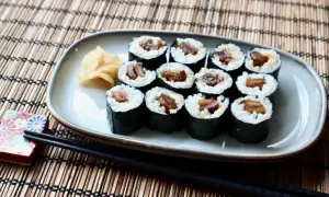 Tykwa sushi, czyli rolki z ryżem i ciemnym nadzieniem, ułożone na talerzu obok imbiru i pałeczek.