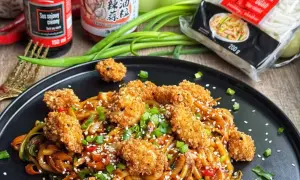 Pyszny udon z kurczakiem i warzywami, posypany sezamem i szczypiorkiem. Obok sos sojowy i opakowanie makaronu udon.