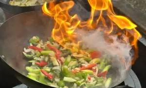 Płomienie otaczają warzywa w woku, tworząc apetyczny stir fry. W tle widać drugie danie.