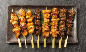 Różnorodne szaszłyki yakitori z kurczakiem i warzywami, polane słodkim sosem, ułożone na ciemnej tacy.