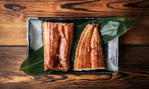 Dwa kawałki lśniącego, glazurowanego unagi na liściu, podane na kamiennej tacy na drewnianym tle.