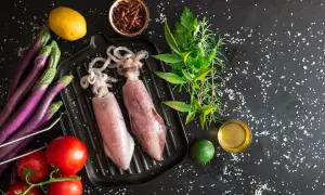 Dwa surowe kalmary na grillu, obok pomidorów, bakłażanów, cytryny, limonki i ziół. Idealne składniki, by dowiedzieć się, jak jeść kalmary.