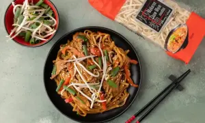 Dania kuchni azjatyckiej: makaron chow mein z warzywami i kiełkami fasoli mung, obok paczka makaronu i pałeczki.