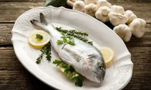 Dorada, co to za ryba? Świeża ryba na talerzu z cytryną i ziołami, obok główki czosnku.