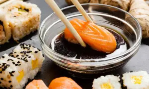Paluszki zanurzają kawałek łososia w sosie sojowym bezglutenowym. Obok sushi i imbir.