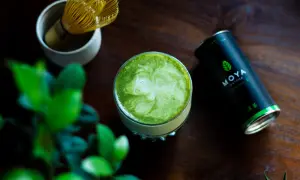 Szklanka zielonego matcha z pianką, obok puszka MOYA Matcha i bambusowa miotełka.