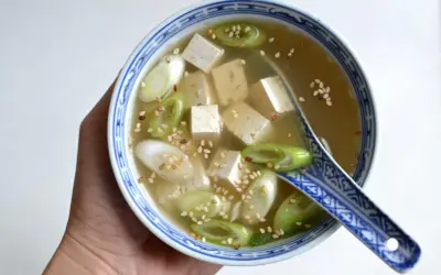 Miseczka z bulionem miso, kostkami tofu, zieloną cebulką i sezamem, trzymana w dłoni.