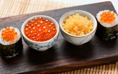 Dwa sushi z ikrą łososia i miseczki z ikrą masago i ikrą łososia na drewnianej desce.