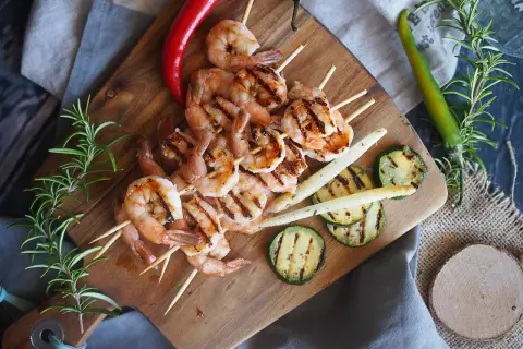 Grillowane krewetki po angielsku na szpadkach, podane z grillowaną cukinią, szparagami i papryczkami chili na drewnianej desce.