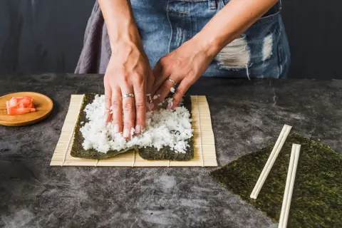 Dłonie rozprowadzają ryż haruka na nori, przygotowując się do zwinięcia sushi. Obok leżą pałeczki i marynowany imbir.
