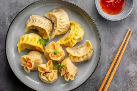 Talerz pełen różnorodnych pierożków dim sum, ozdobionych natką pietruszki, z miseczką sosu i pałeczkami.
