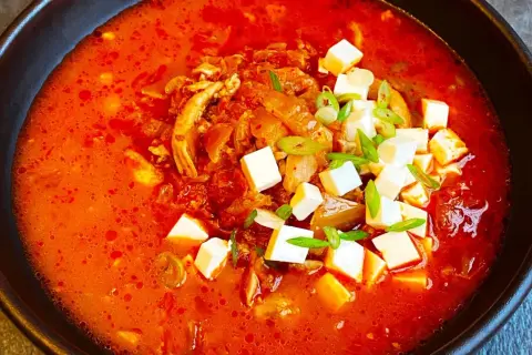 Pikantna zupa kimchi z kurczakiem i kostkami tofu, udekorowana szczypiorkiem.