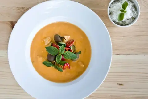 Aksamitny **thai red curry** z kurczakiem, cukinią i pomidorkami, udekorowany bazylią. Obok miseczka ryżu.
