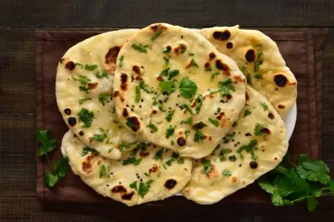 Stos chlebków naan, posypanych kolendrą i czarnuszką, z widocznymi pęcherzykami powietrza. Idealne do curry, ale warto sprawdzić chlebek naan kcal.