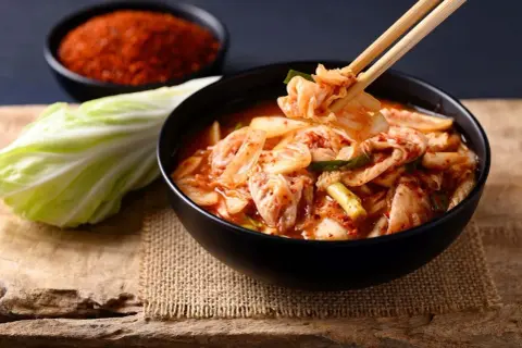 Miseczka z kimchi, pałeczki podnoszące kawałek kapusty, liść kapusty i miseczka z przyprawą.