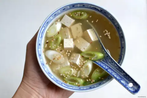 Miseczka z bulionem miso, kostkami tofu, zieloną cebulką i sezamem, trzymana w dłoni.