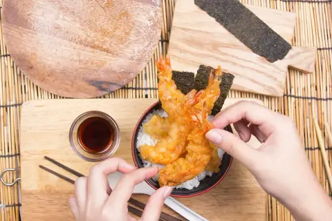 Ręce sięgają po chrupiące krewetki w panierce tempura, ułożone na ryżu z nori.