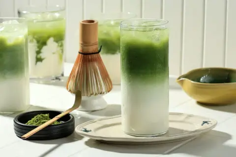 Orzeźwiające matcha latte z lodem, idealne dla mam karmiących piersią. Obok bambusowa miotełka i łyżeczka do przygotowania napoju.