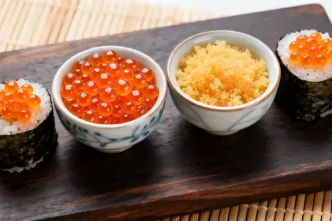 Dwa sushi z ikrą łososia i miseczki z ikrą masago i ikrą łososia na drewnianej desce.