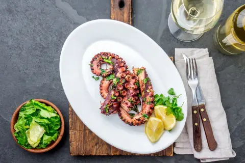 Grillowane macki ośmiornicy z cytryną i ziołami, podane na białym talerzu z sałatką i kieliszkiem białego wina.