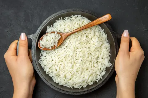 Dwie dłonie trzymają okrągłą patelnię z parującym ryżem basmati. Drewniana łyżka zanurzona w ryżu.