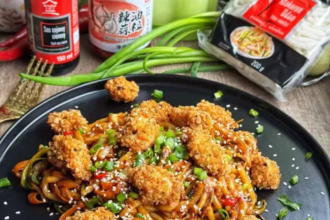 Pyszny udon z kurczakiem i warzywami, posypany sezamem i szczypiorkiem. Obok sos sojowy i opakowanie makaronu udon.
