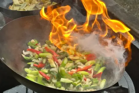 Płomienie otaczają warzywa w woku, tworząc apetyczny stir fry. W tle widać drugie danie.
