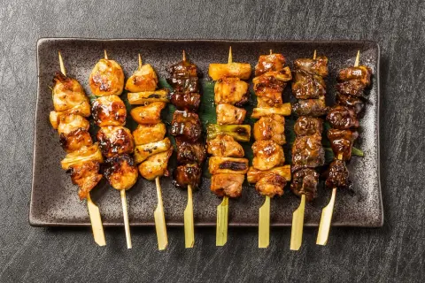 Różnorodne szaszłyki yakitori z kurczakiem i warzywami, polane słodkim sosem, ułożone na ciemnej tacy.