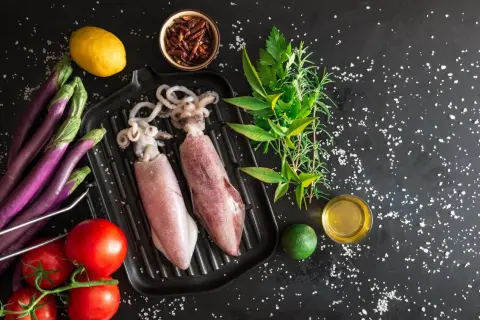 Dwa surowe kalmary na grillu, obok pomidorów, bakłażanów, cytryny, limonki i ziół. Idealne składniki, by dowiedzieć się, jak jeść kalmary.
