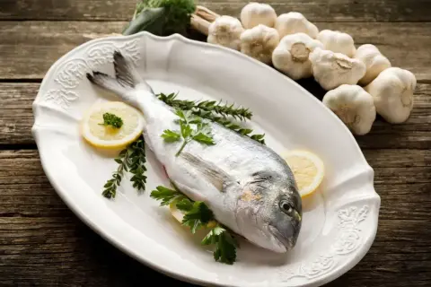 Dorada, co to za ryba? Świeża ryba na talerzu z cytryną i ziołami, obok główki czosnku.