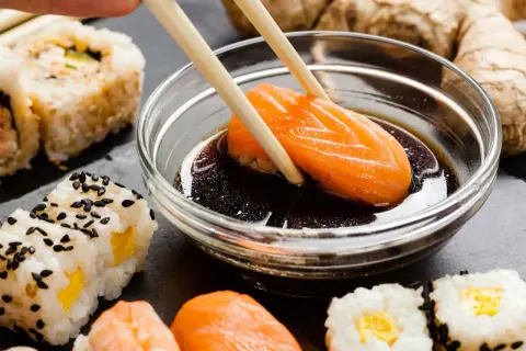 Paluszki zanurzają kawałek łososia w sosie sojowym bezglutenowym. Obok sushi i imbir.