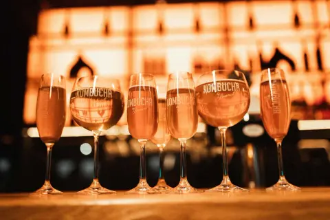 Sześć kieliszków z napojem kombucha na barze, w tle rozmyte światła.