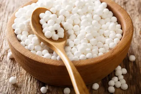Drewniana miska pełna białych kuleczek, które wyglądają jak makaron z manioku. Drewniana łyżka zanurzona w kuleczkach.