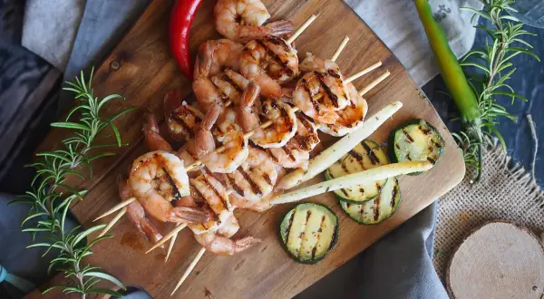 Grillowane krewetki po angielsku na szpadkach, podane z grillowaną cukinią, szparagami i papryczkami chili na drewnianej desce.