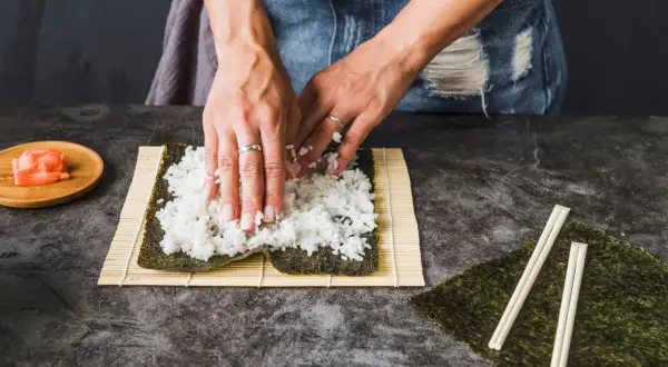 Dłonie rozprowadzają ryż haruka na nori, przygotowując się do zwinięcia sushi. Obok leżą pałeczki i marynowany imbir.