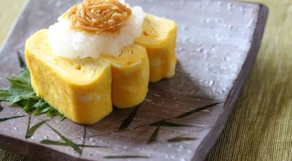 Tamago co to? Japoński omlet tamagoyaki, zwinięty w rolki, podany z tartą rzodkwią i grzybami enoki na talerzu z bambusowym wzorem.