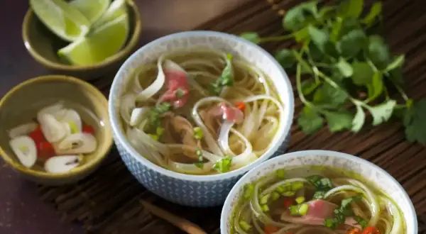 Dwie miseczki aromatycznej zupy pho, z cienkim makaronem ryżowym, plastrami wołowiny i świeżą kolendrą. Obok miseczki z limonką i marynowanym imbirem. To przepis na oryginalną zupę pho.