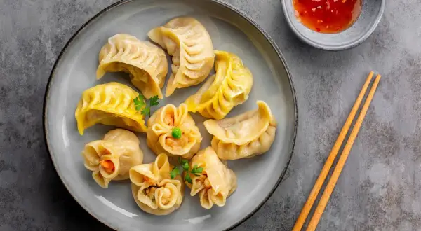 Talerz pełen różnorodnych pierożków dim sum, ozdobionych natką pietruszki, z miseczką sosu i pałeczkami.