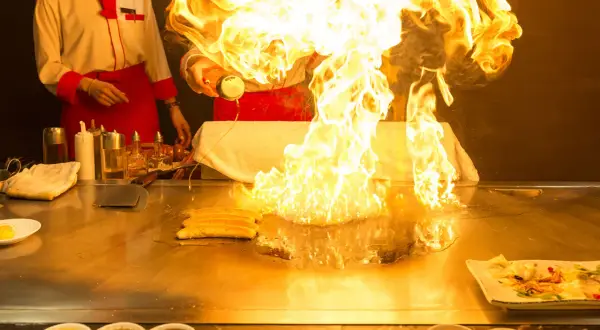 Szef kuchni tworzy spektakularny pokaz ognia podczas przygotowywania yakiniku. Płomienie unoszą się nad płytą grillową, a obok leżą kawałki mięsa.