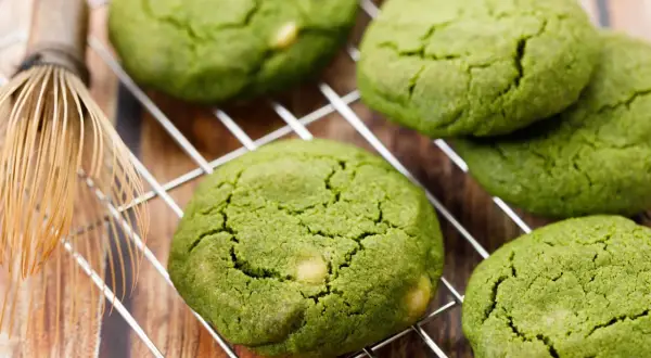Zielone, japońskie ciasteczka matcha z kawałkami białej czekolady na metalowej kratce, obok bambusowej miotełki do herbaty.