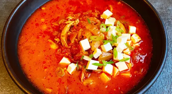 Pikantna zupa kimchi z kurczakiem i kostkami tofu, udekorowana szczypiorkiem.
