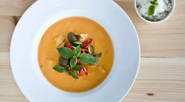 Aksamitny **thai red curry** z kurczakiem, cukinią i pomidorkami, udekorowany bazylią. Obok miseczka ryżu.