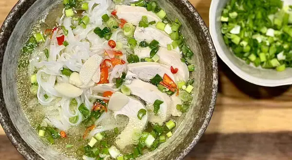 Aromatyczna zupa pho ga z kawałkami kurczaka, makaronem ryżowym, szczypiorkiem i papryczką chili. Obok miseczka z posiekanym szczypiorkiem.